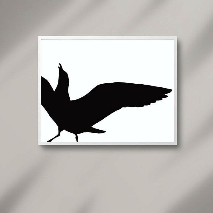 Printable wall art of the black silhouette of a seagull set against a white background, displayed in a white frame on a grey wall.
