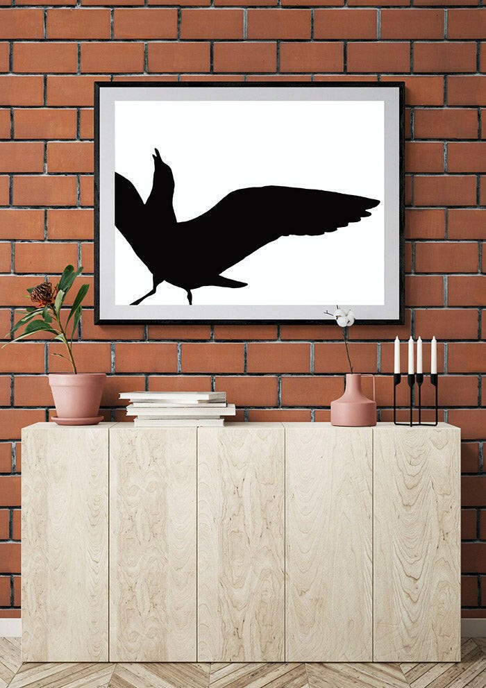 Framed black silhouette of a seagull on a white background against a brick wall with a wooden cabinet below.